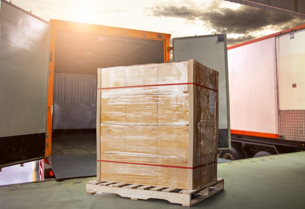 Palé de cajas retractilado con fleje en muelle de carga junto a camiones, listo para traslado a terminal